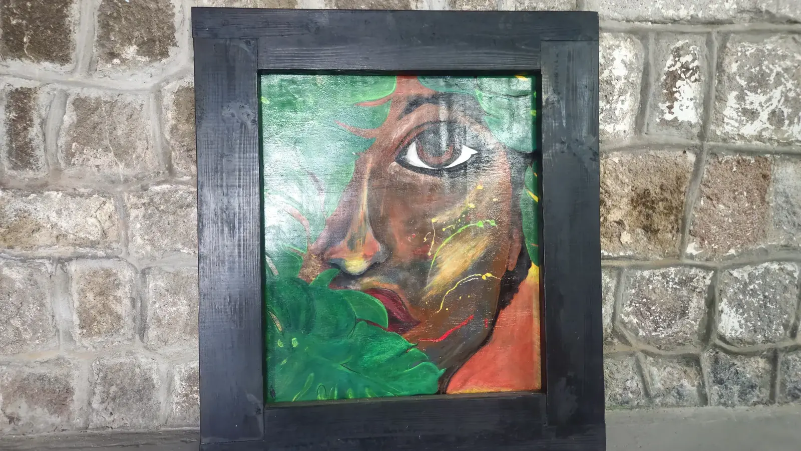 A framed painting of a face half-hidden behind green leaves, displayed on the gallery's stone wall
